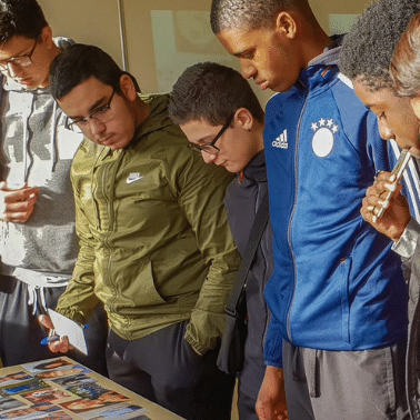 Young men, various ages and ethnicities, peer down at a table with images