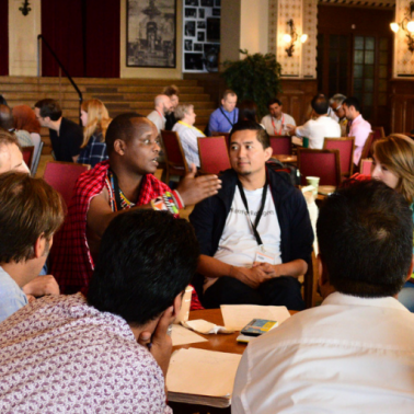 Black man with African shawl reaches out towards a group of mixed gendered people during a small group discussion