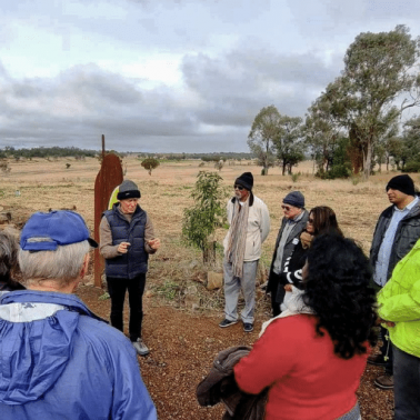 White male speaking to semi-circle of white and indigenous Australians outdoors