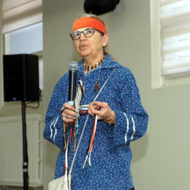 Canadian Trustbuilding panel speakers and outreach event Indigenous Canadian woman, wearing blue tunic and orange headband, speaks before a crowd